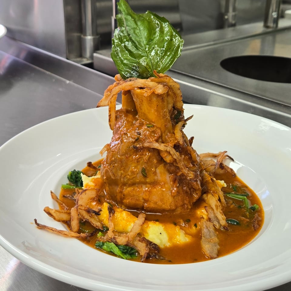 Braised osso buco with mashed potatoes, spinach, crispy onions, and a large basil leaf.